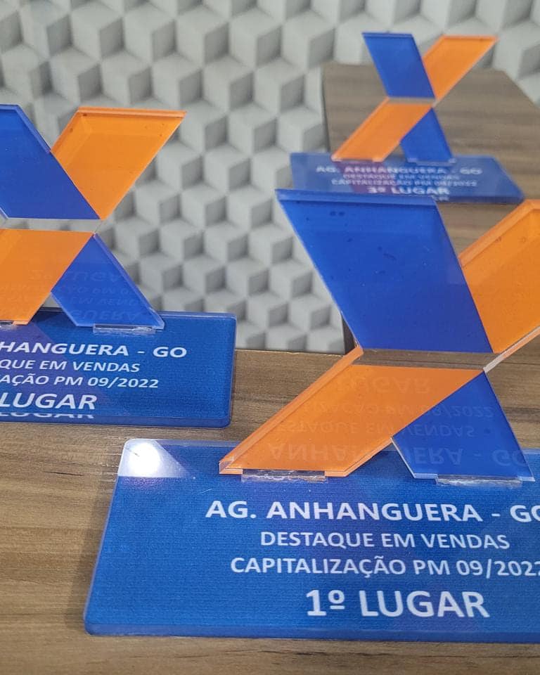 Troféus de destaque em vendas da Caixa, em acrílico laranja e azul no formato do X da marca, com a placa impressa na base