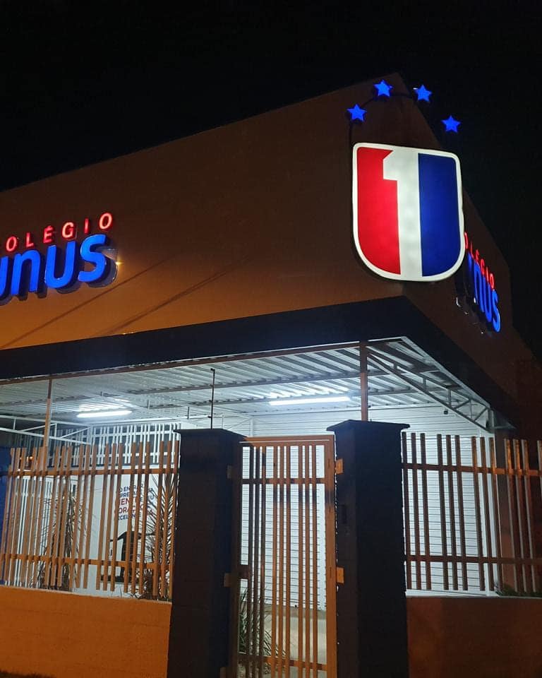 Fachada do Colégio Unus à noite, com o símbolo e as letras caixa iluminadas sobre o volume em ACM