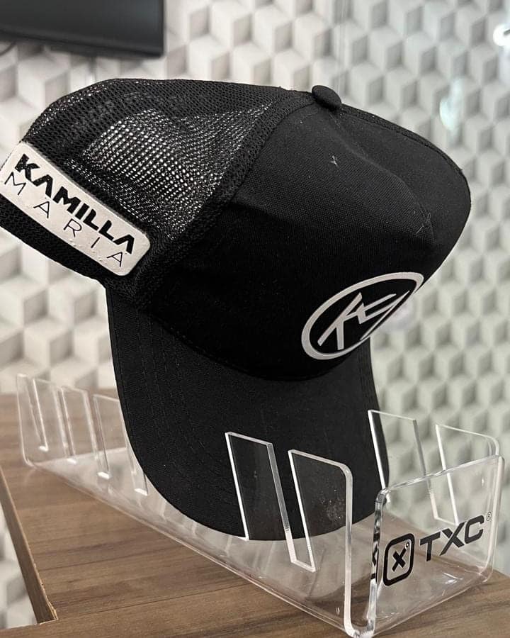 Porta-boné em acrílico transparente com a marca TXC aplicada na base, exibindo um boné TXC com etiqueta personalizada de Kamilla Maria