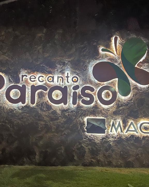 Letra caixa do Recanto Paraíso iluminada por trás à noite, sobre o muro de pedra