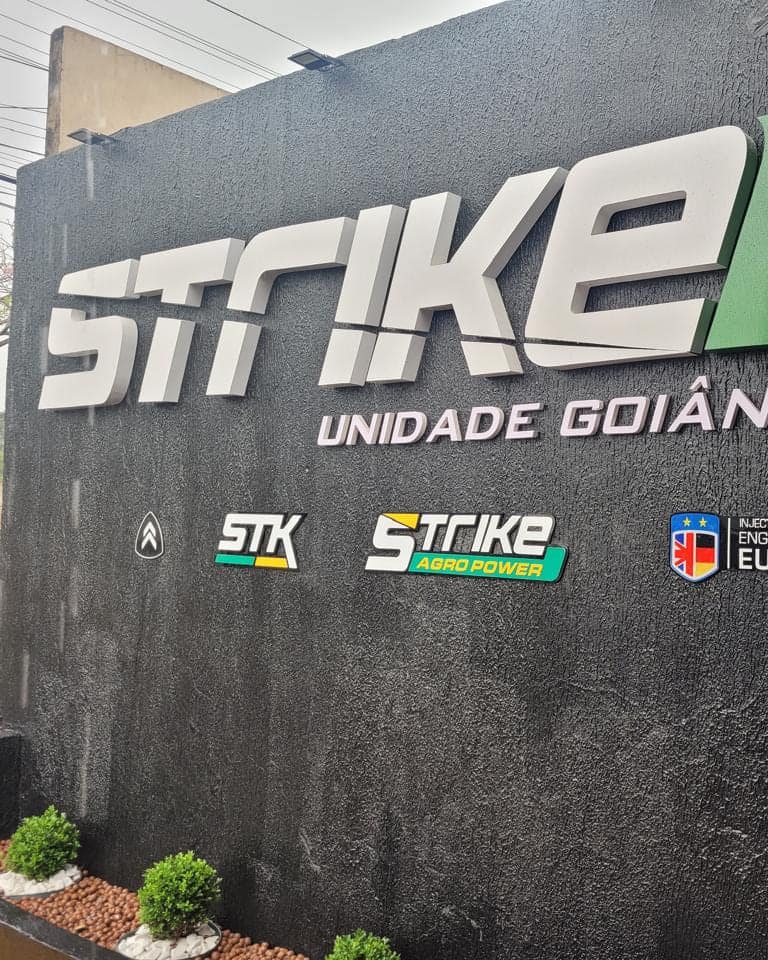 Letra caixa metálica da Strike aplicada sobre muro texturizado preto, com as submarcas alinhadas abaixo