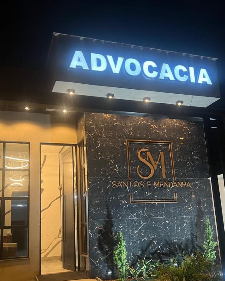 Entrada do escritório Santos e Mendonça Advocacia à noite, com a palavra Advocacia iluminada no alto e o monograma em metal sobre parede de pedra preta