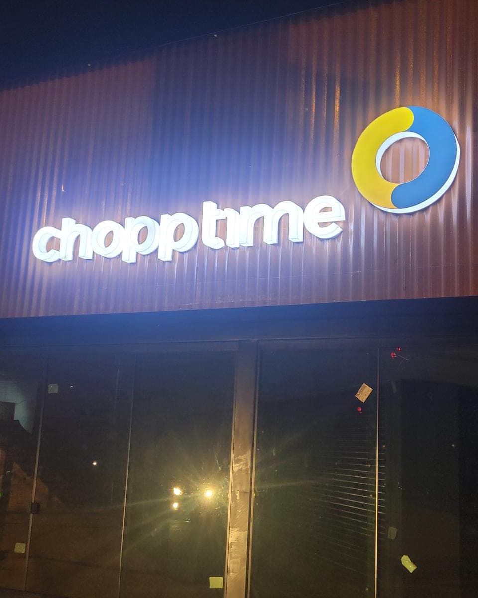 Letreiro da Chopp Time aceso à noite, com letras brancas iluminadas e o símbolo em amarelo e azul sobre fachada de chapa ondulada