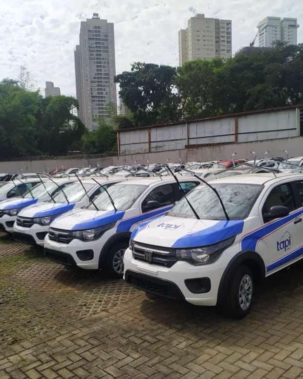 Frota de carros compactos brancos alinhados no pátio, todos com a mesma adesivagem azul nas portas e no capô