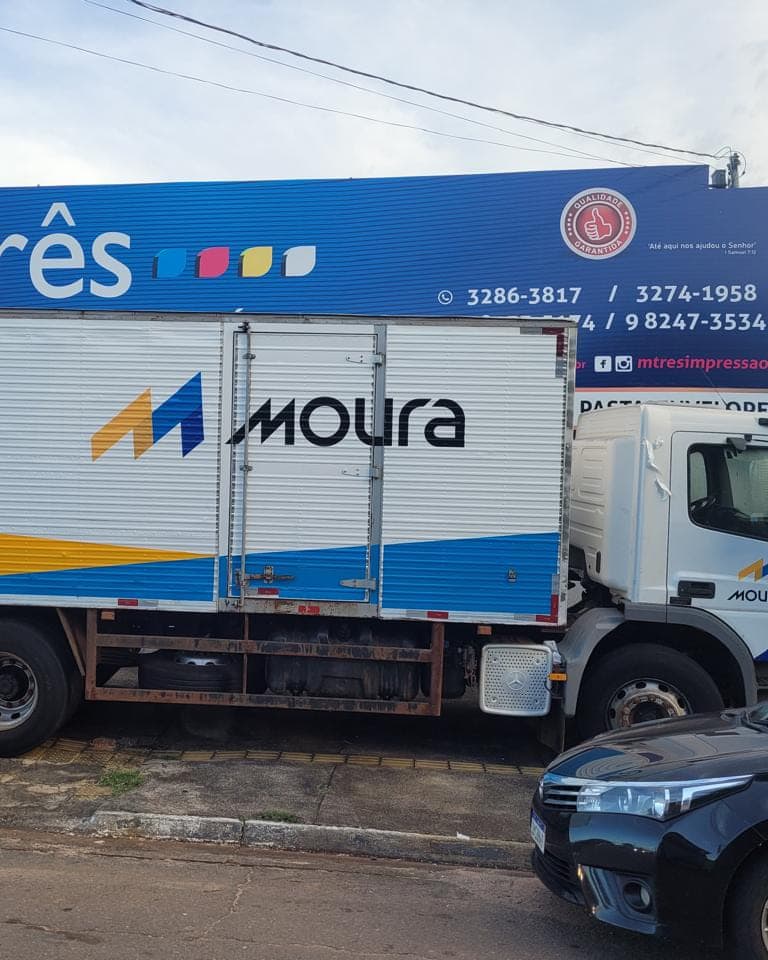 Baú de caminhão da Moura adesivado em branco, azul e amarelo, com a marca e a chamada de venda na lateral