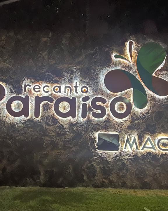 Letra caixa do Recanto Paraíso iluminada por trás à noite, sobre o muro de pedra