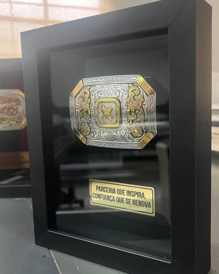 Quadro preto com fivela de cinto em relevo e placa dourada gravada, produzido para a TXC