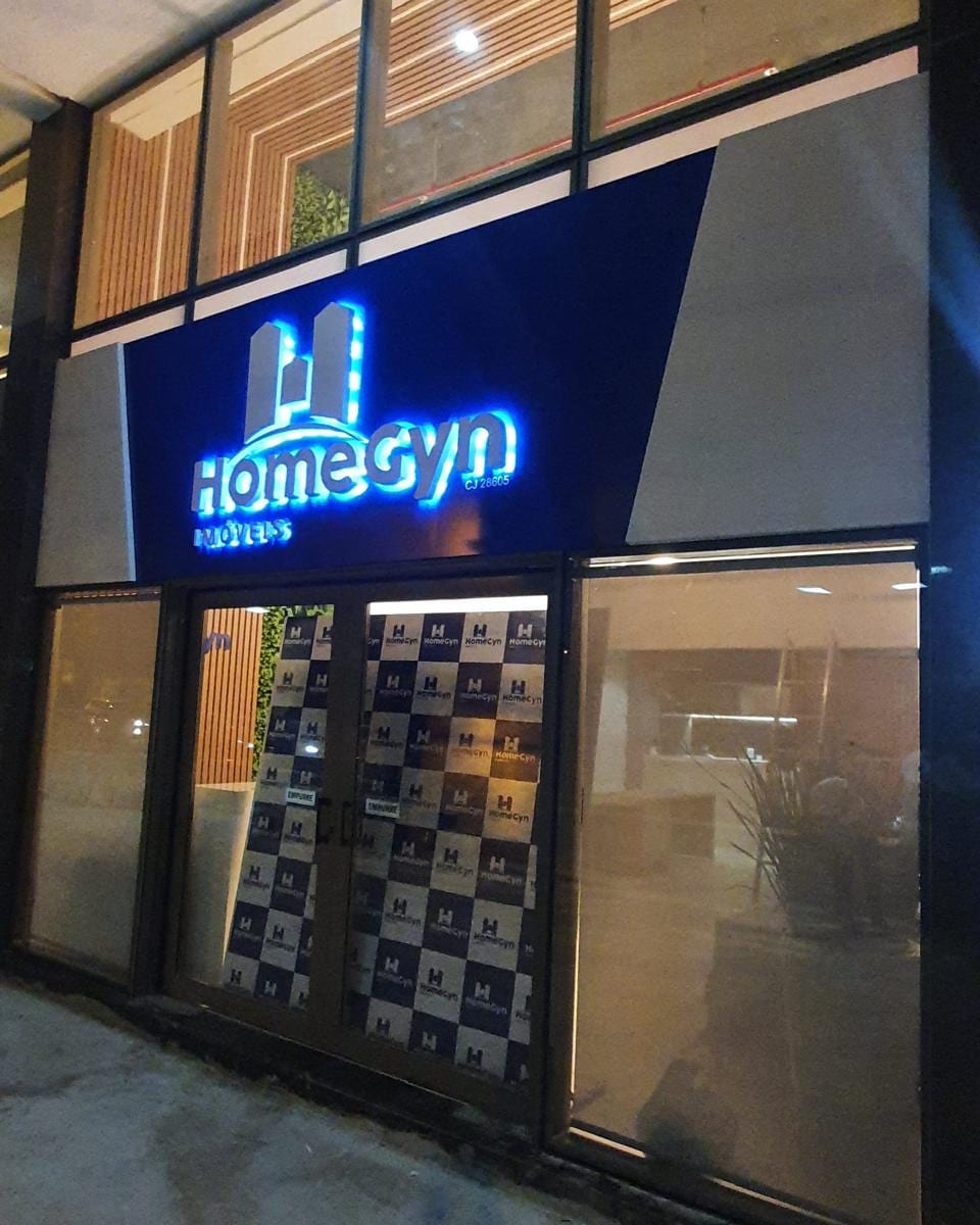 Fachada da HomeGyn Imóveis à noite, com o letreiro em letra caixa iluminada em azul sobre painel escuro e a vitrine adesivada com o padrão da marca